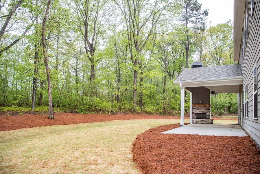 Backyard with covered patio, stone fireplace, and grassy area surrounded by trees and mulch.