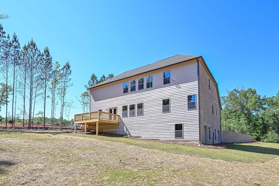 Rear view of a two-story house with a wooden deck on a grassy hillside under a blue sky.