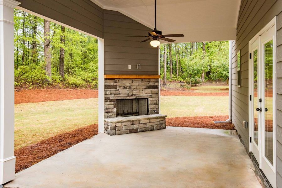 Covered patio with stone fireplace, ceiling fan, and French doors overlooking a grassy yard and trees.