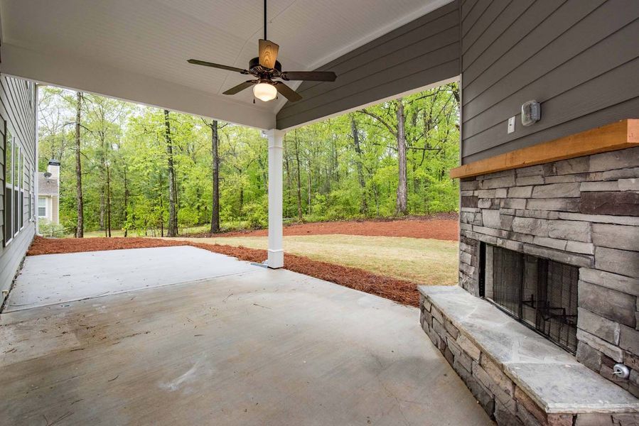 Covered patio with fireplace, ceiling fan, and view of trees. Gray stone, wood, and concrete.