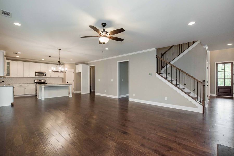 Open-plan living area with kitchen, stairs, and dark wood floors. Light walls, white cabinets, and a ceiling fan.
