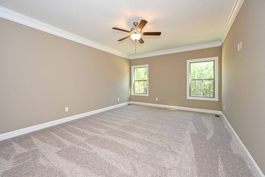 Empty bedroom with beige walls, light carpet, two windows, and a ceiling fan.