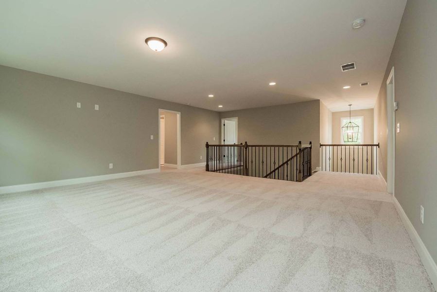 Empty, carpeted loft space with gray walls, stair railing, and overhead lights.