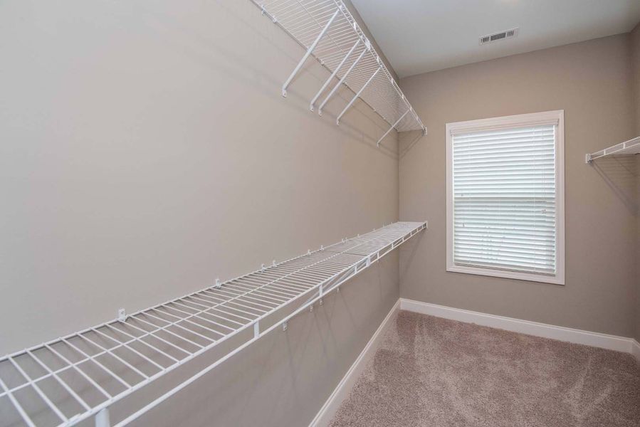 Empty walk-in closet with white wire shelving and a window with closed blinds. Beige walls and carpet.