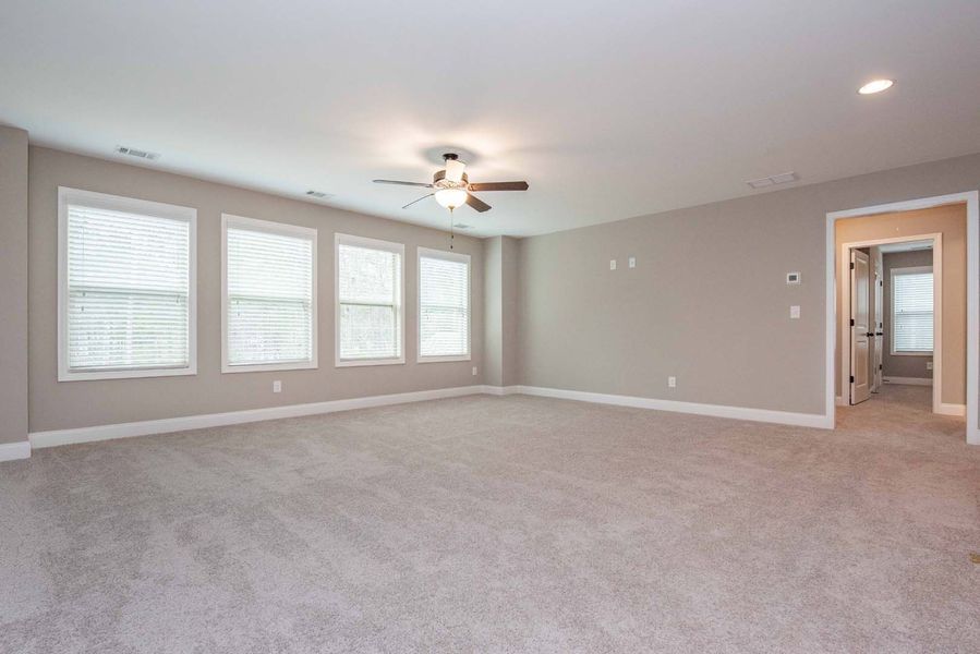Empty, spacious room with light gray walls and carpet, windows with blinds, and a ceiling fan.