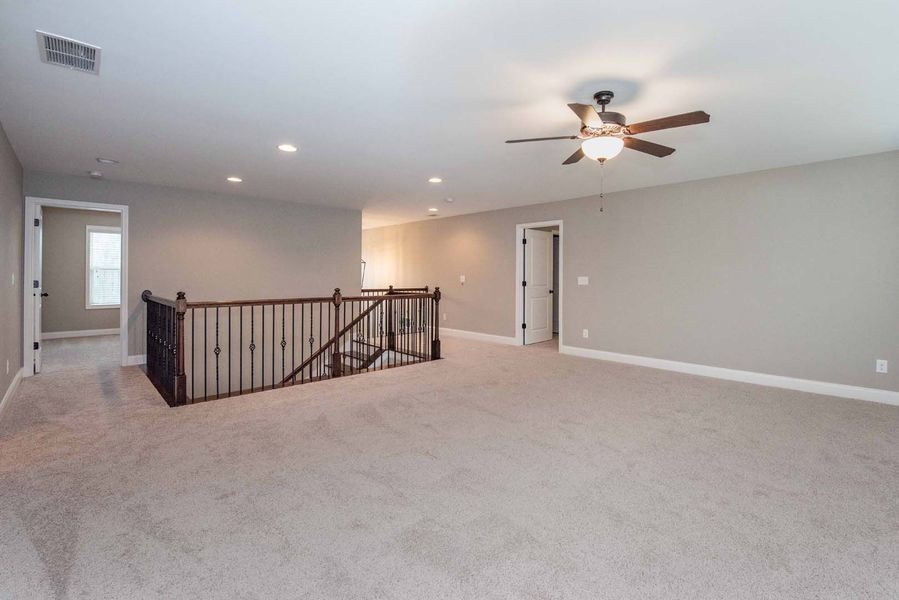 Open, carpeted loft area with a dark banister and ceiling fan. Neutral walls, doors, and a hallway.