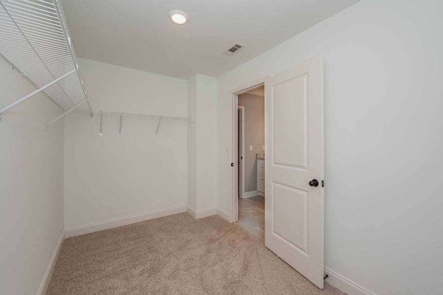 Empty walk-in closet with wire shelving, open door to a bathroom, and tan carpet.