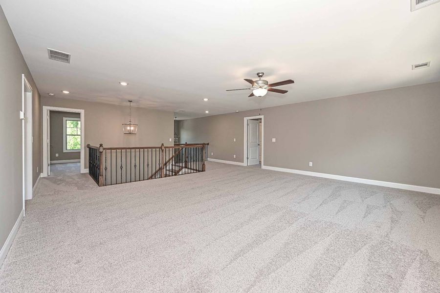 Spacious, neutral-toned carpeted loft with railing, stairs, and ceiling fan.