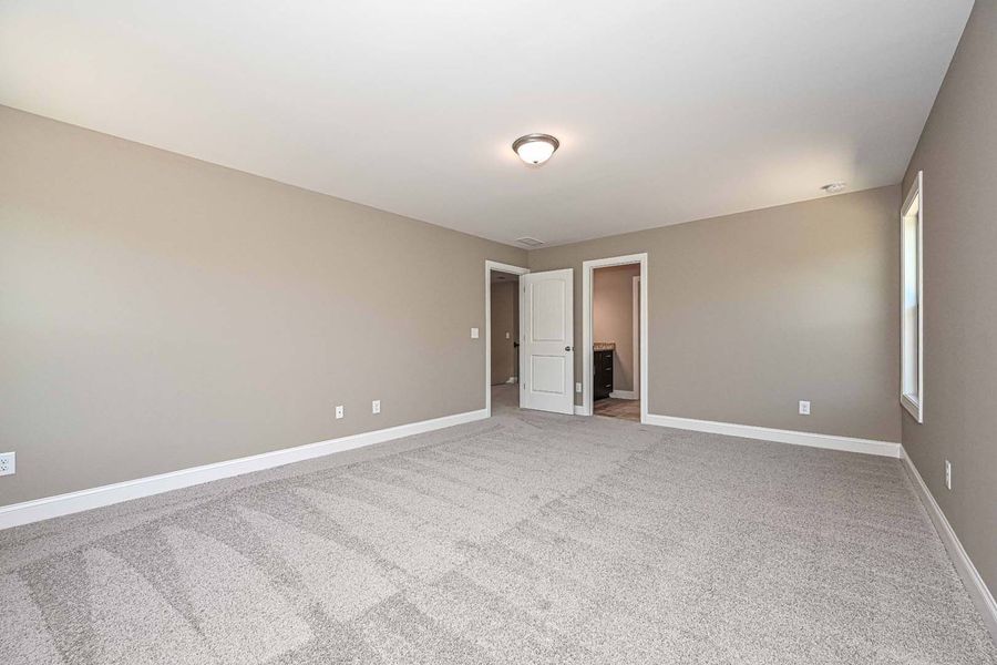 Empty bedroom with beige walls, gray carpet, and white trim. Two doors and a window are visible.