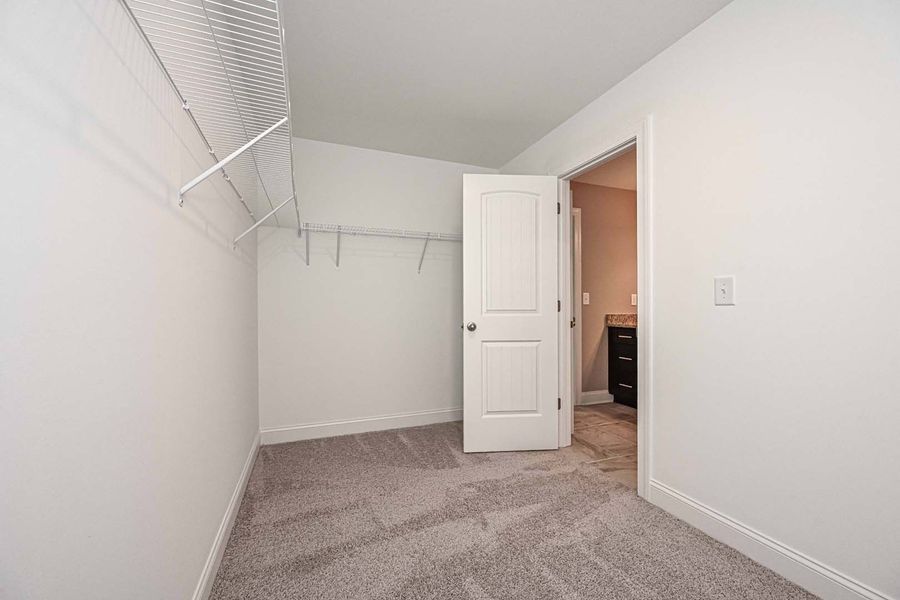 Empty walk-in closet with white walls, wire shelves, and a door leading to a bathroom. Carpeted floor.