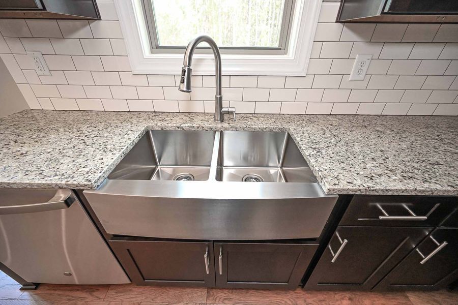 Stainless steel kitchen sink with granite countertop, white subway tile backsplash, and dark cabinets.