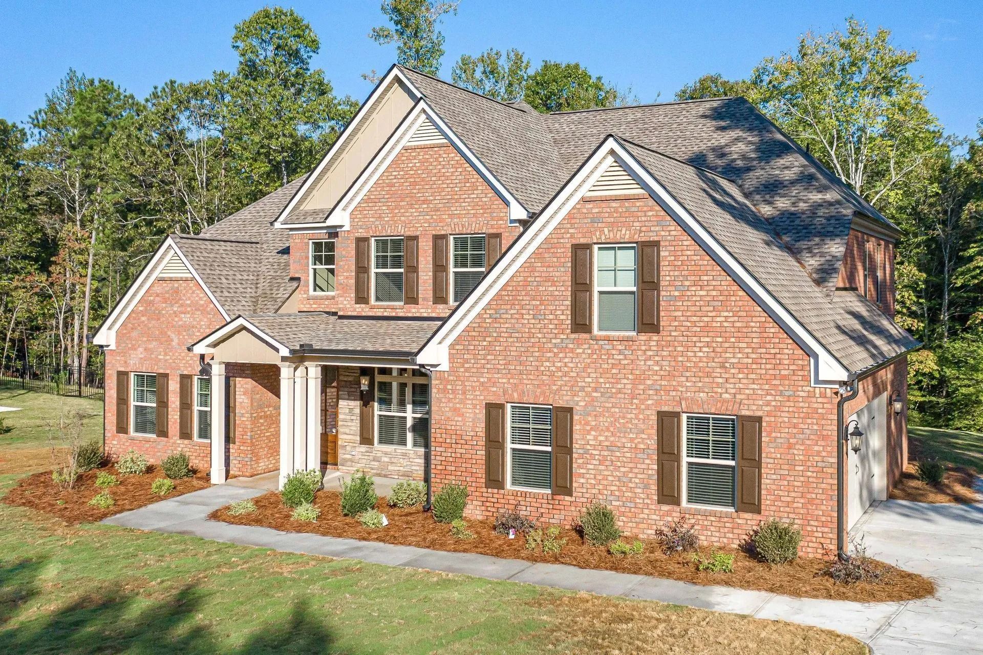 Brick two-story house with brown shutters and roof, surrounded by trees and landscaping on a sunny day.