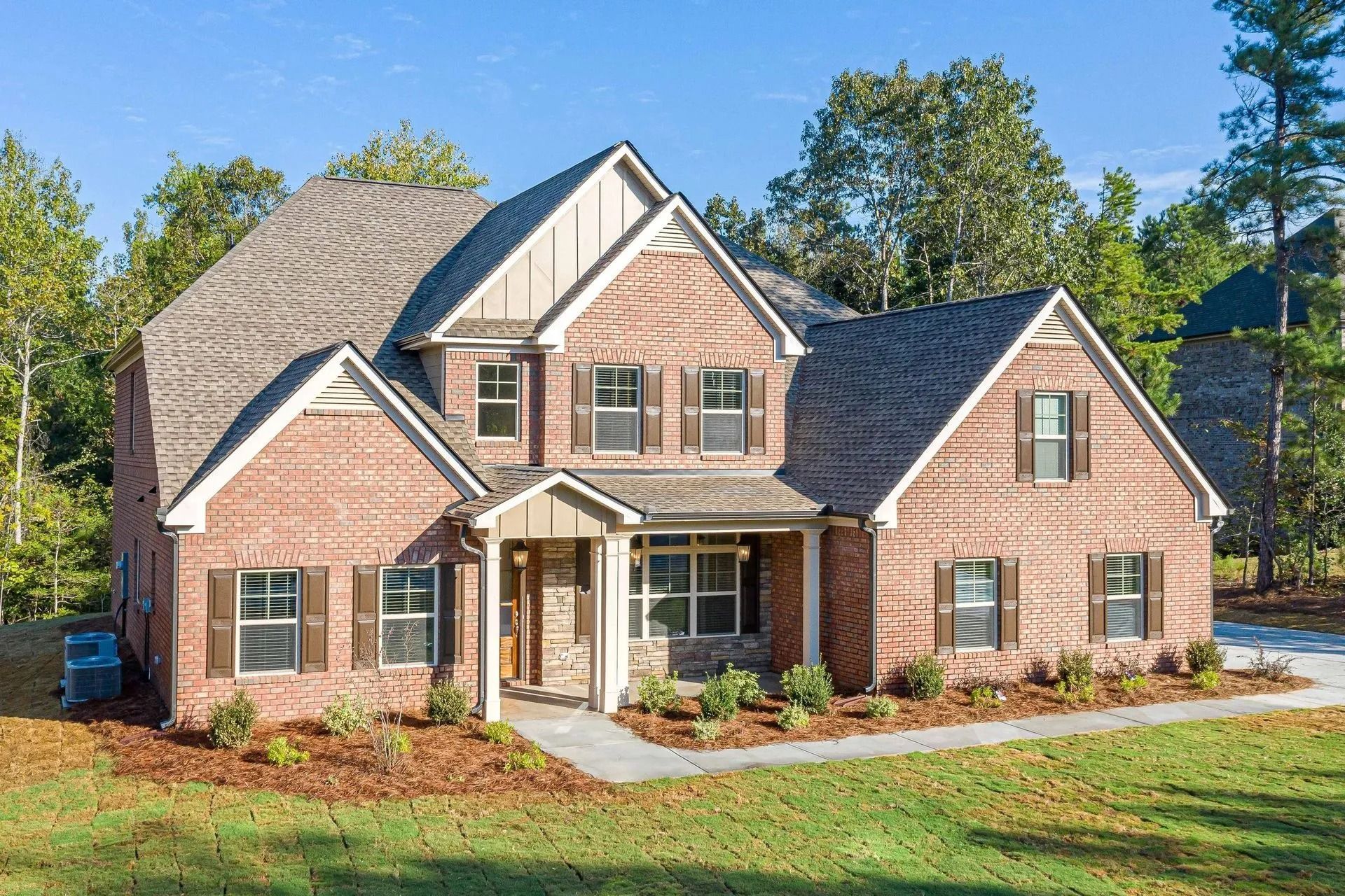 Two-story brick house with brown shutters and roof, green lawn, trees, and blue sky.