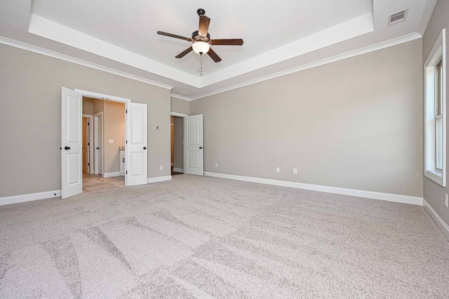 Empty bedroom with beige walls, carpet, ceiling fan, and white trim around doors and windows.