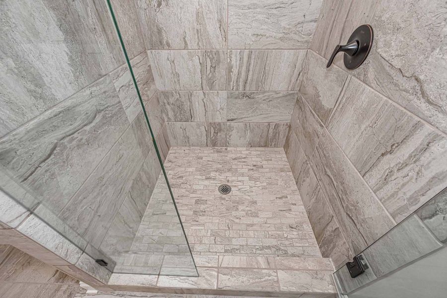 Shower interior with light grey tile and a glass door. Black shower head.