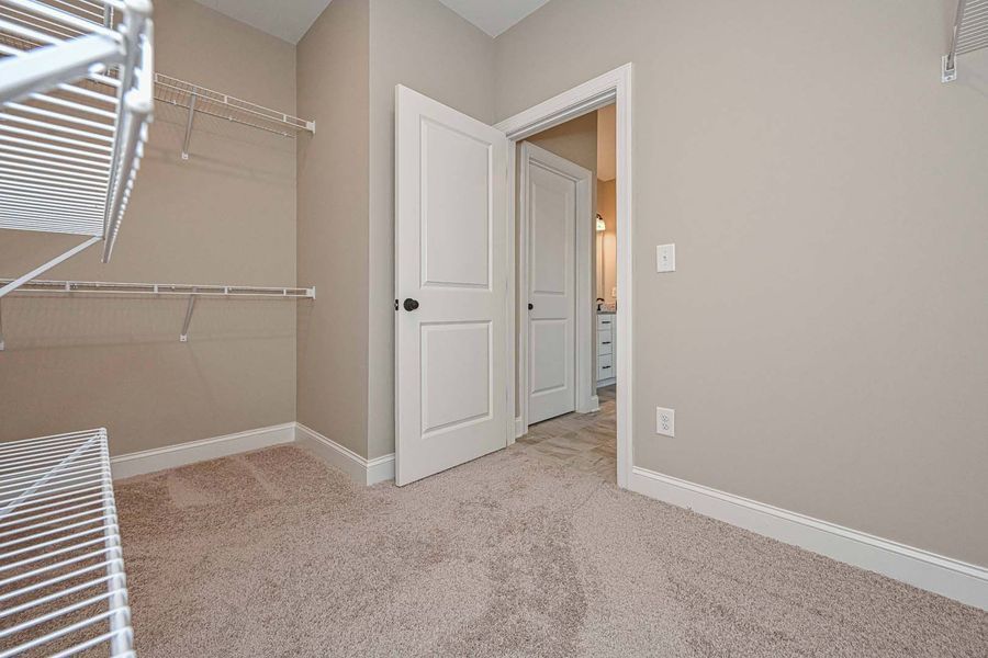 Empty walk-in closet with beige carpet, wire shelving, and an open door leading to a bathroom.