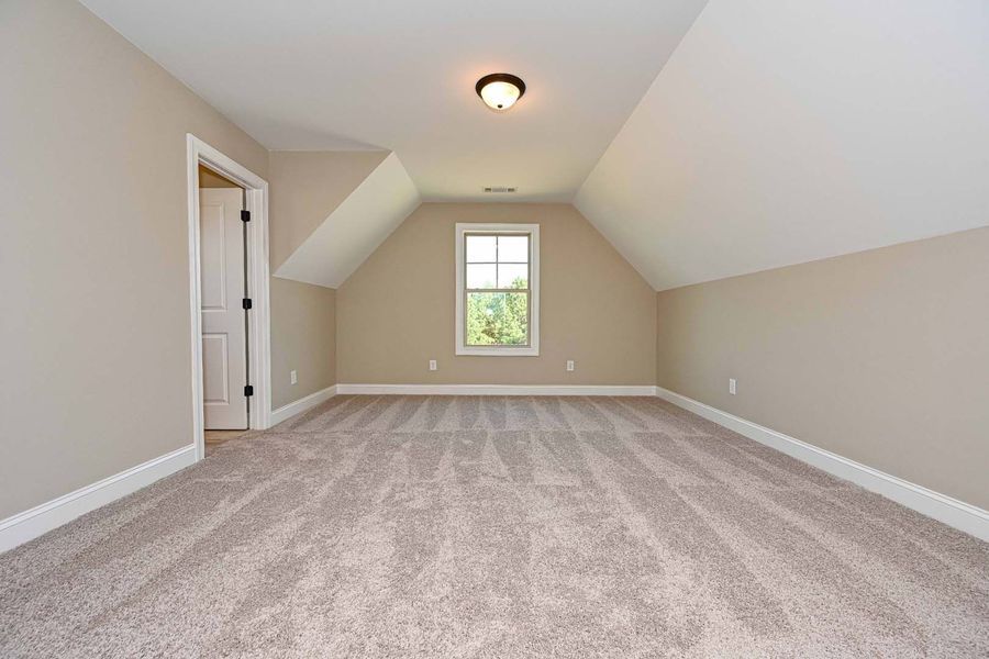 Empty, beige-walled attic room with carpet, white trim, a small window, and a door.