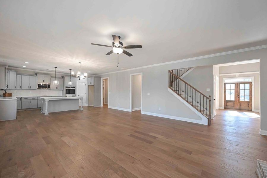 Open-concept living space with kitchen, staircase, and entryway. Light hardwood floors, gray walls, and a ceiling fan.