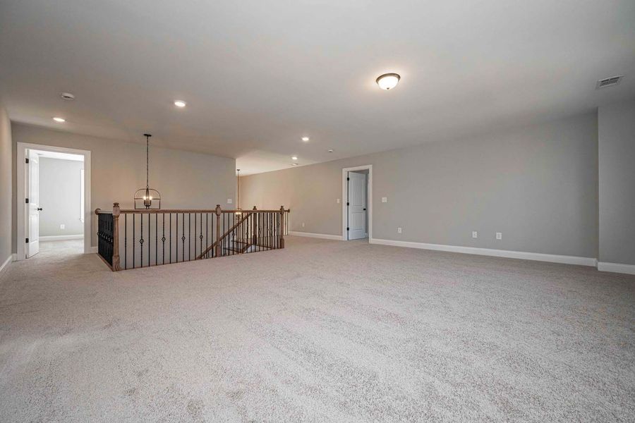Spacious upper-level loft with gray walls, carpet, and a wooden railing. Two doors and recessed lighting are visible.