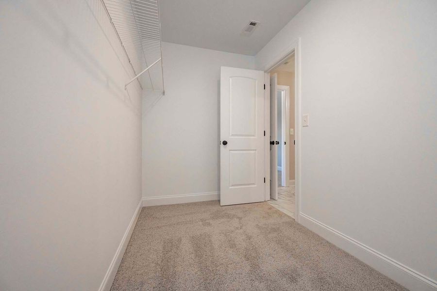 Empty walk-in closet with white walls, gray carpet, and a door leading to another room.
