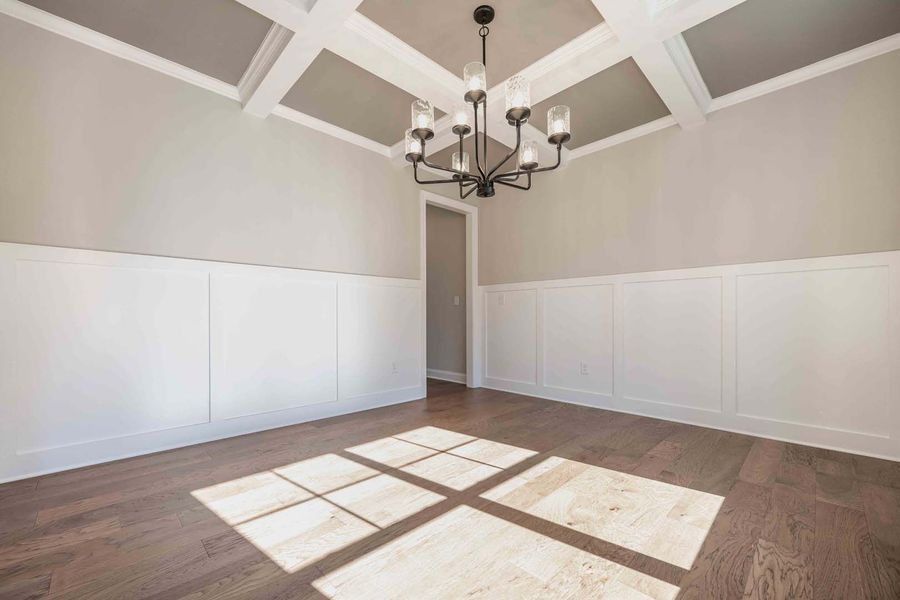 Empty dining room with white wainscoting, coffered ceiling, chandelier, and sunlight on hardwood floor.