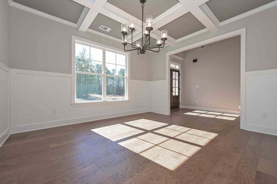 Empty dining room with hardwood floors, white walls, grey ceiling, and chandelier. Sunlight through a window.
