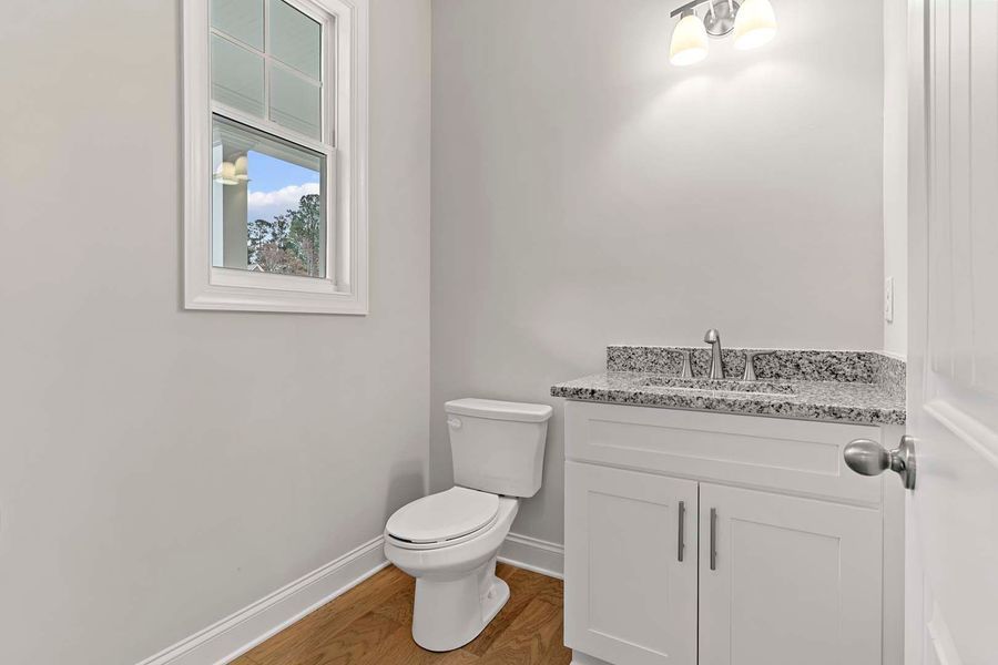 A small, modern bathroom with a white toilet, vanity, and light gray walls; a window on the left.