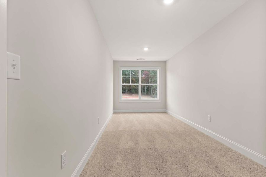 Empty, narrow room with beige carpet, white walls, window at the end, and recessed ceiling light.