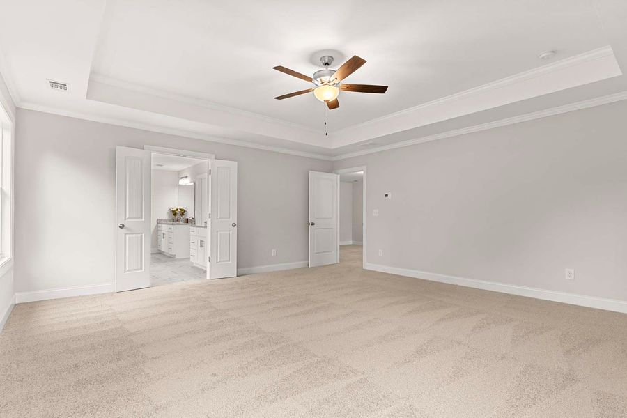 Empty master bedroom with neutral carpet, light walls, and a ceiling fan. Doors lead to the bathroom and hallway.