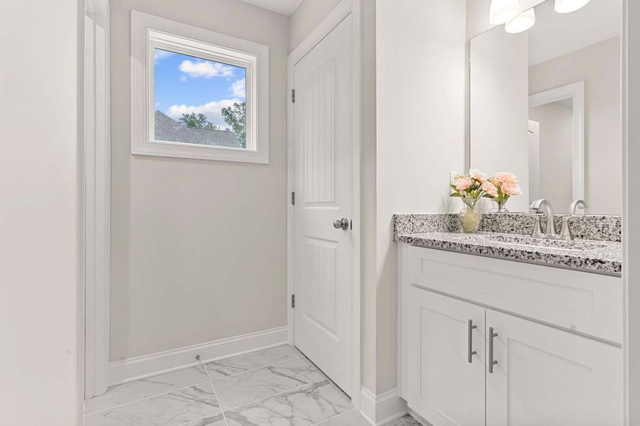 White bathroom with marble tile floor, a vanity with a floral arrangement, and a window with a blue sky view.