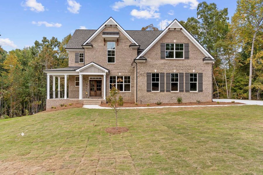 Two-story brick house with a porch, surrounded by grass and trees under a blue sky.