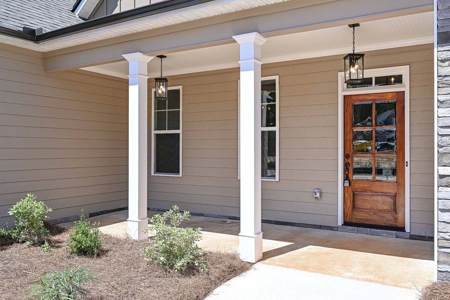 Beige house front porch with white columns, brown door, two windows, and hanging lights.