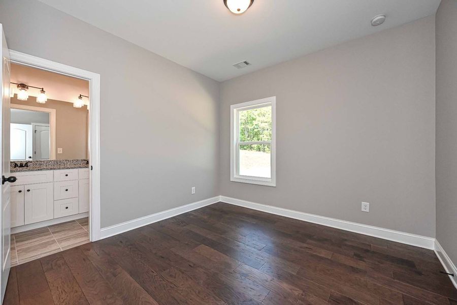 Empty bedroom with dark wood floors, gray walls, a small window, and an open doorway to a bathroom.