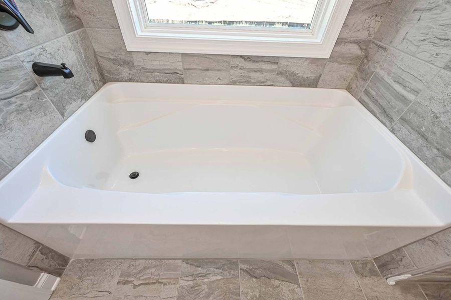 White bathtub in a bathroom with gray tile walls, beneath a window.