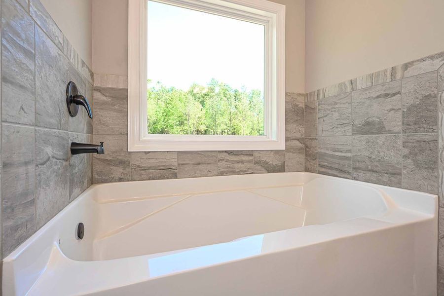 White bathtub under a window with a view of trees, gray tiled walls, and black faucet.