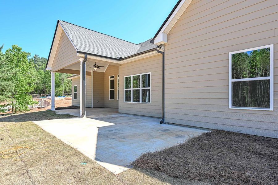 Tan house exterior with a covered patio and large windows, set against a blue sky and greenery.
