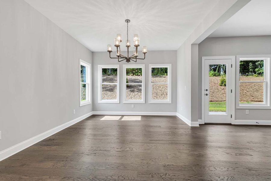 Empty dining room with dark wood floors, light gray walls, and chandelier. Windows overlook greenery.