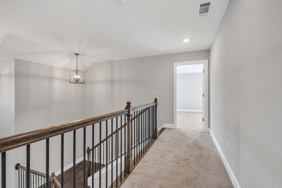 Hallway with carpet, railings, and a light fixture, leading to an open doorway and stairs below.