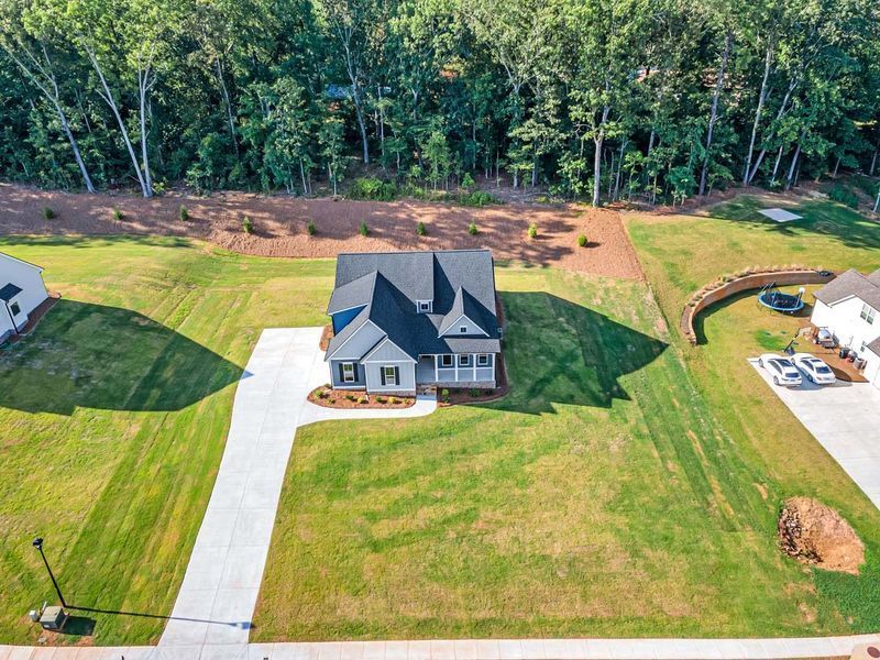 Aerial view of a light blue house with a white porch and black roof, set on a green lawn near a forest.