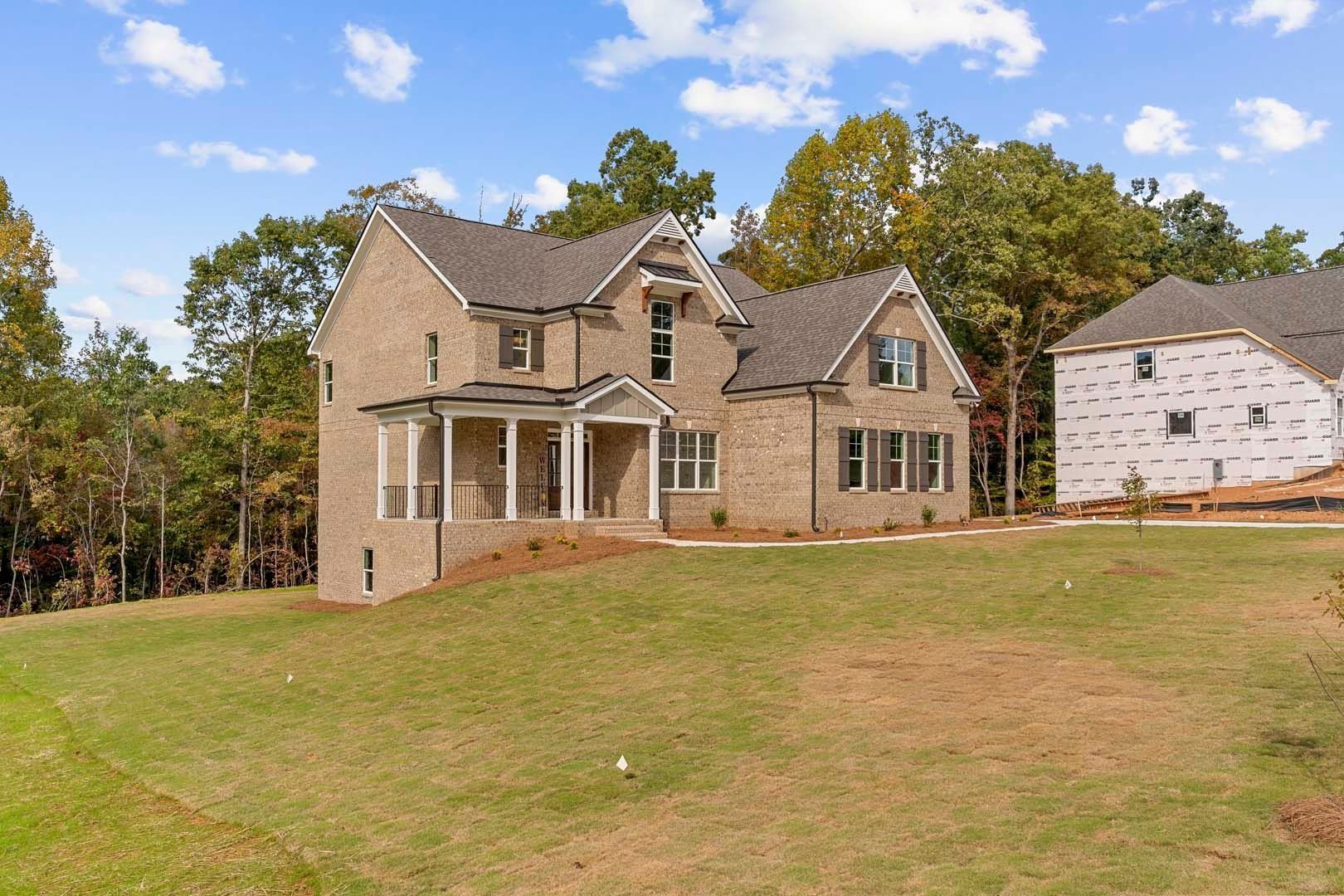 Two-story brick house with a porch on a grassy lot; trees in the background, another house under construction.