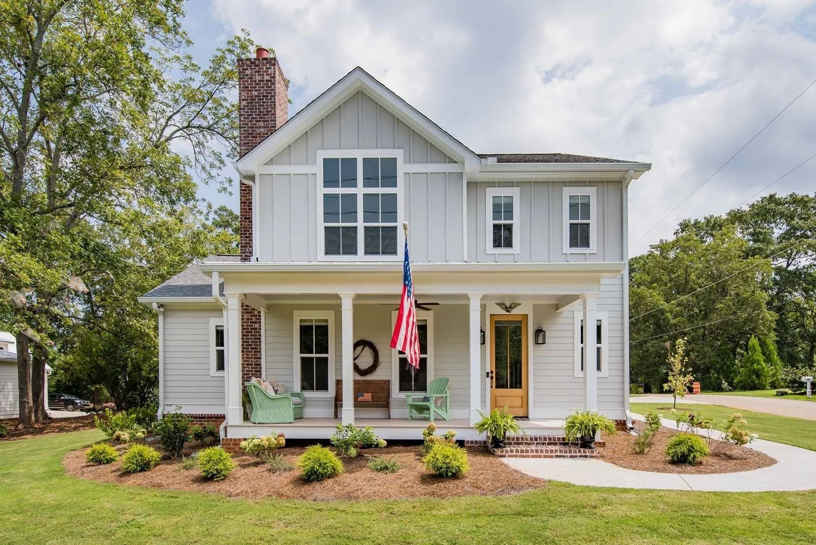 Two-story light gray house with a porch and American flag. Brick chimney and landscaping visible.