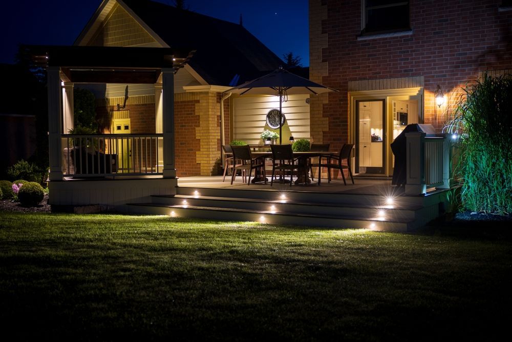 Nighttime view of a home's illuminated patio and steps, with outdoor seating and lush landscaping.
