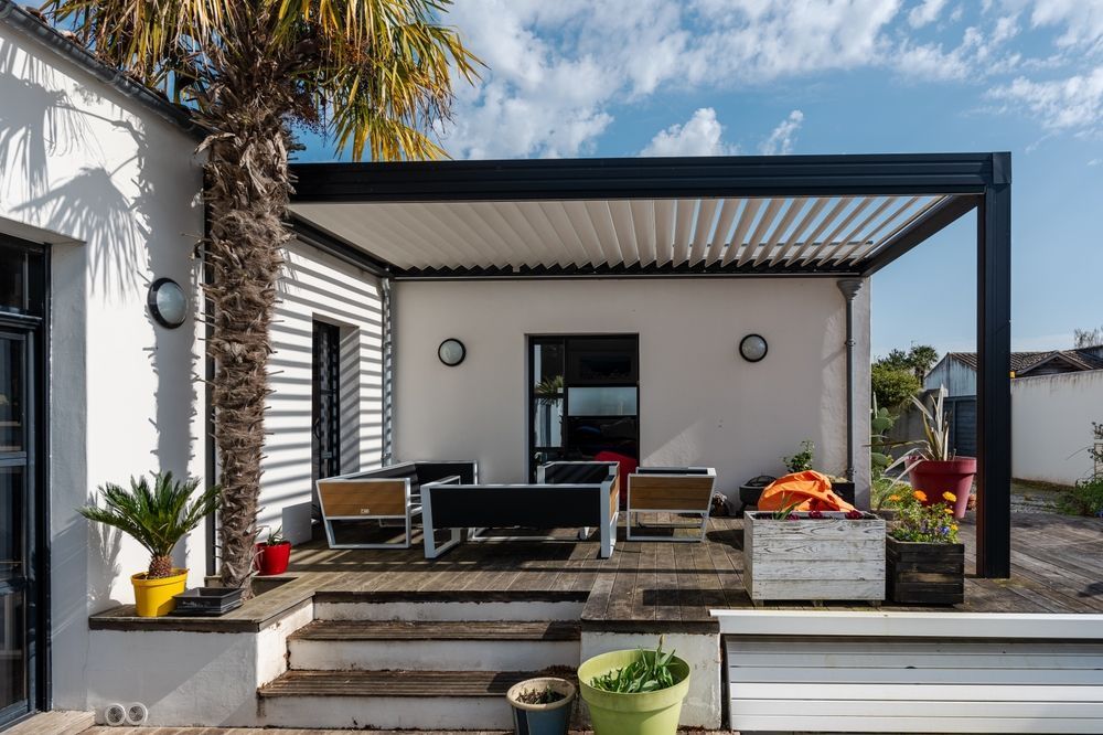 Patio with black pergola, white walls, wooden deck, furniture, and palm tree.