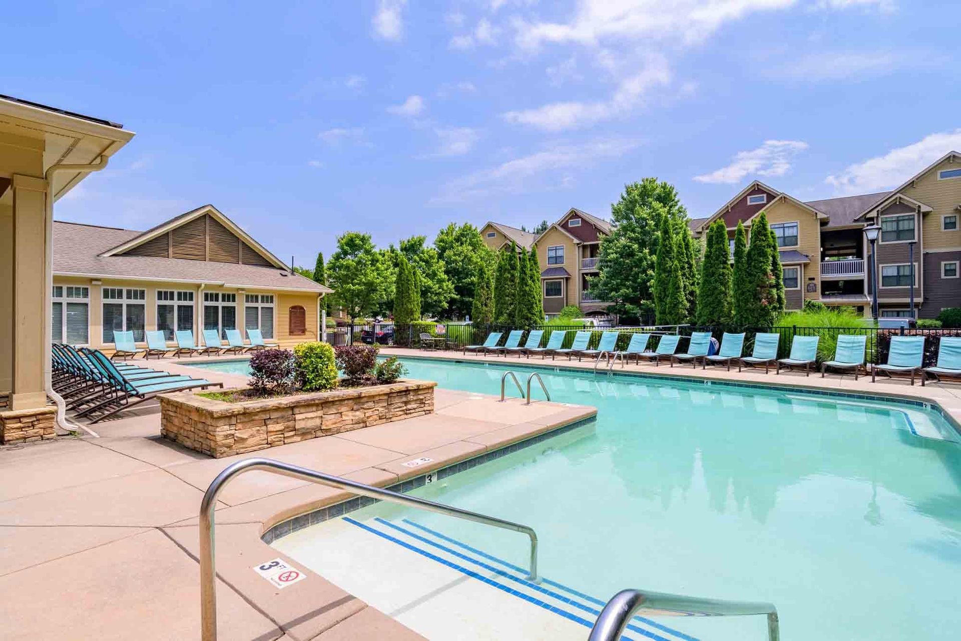 Swimming pool with lounge chairs, buildings in the background, blue sky.