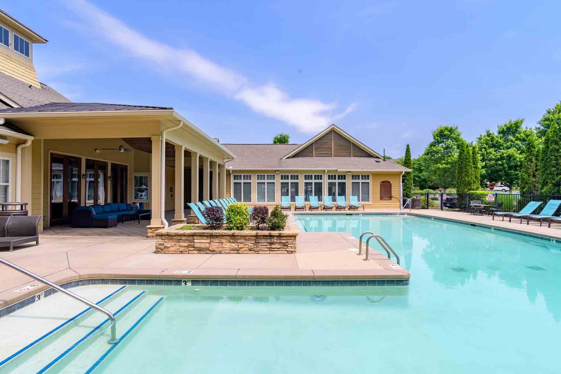 Pool area with turquoise water, beige buildings, and blue lounge chairs on a sunny day.