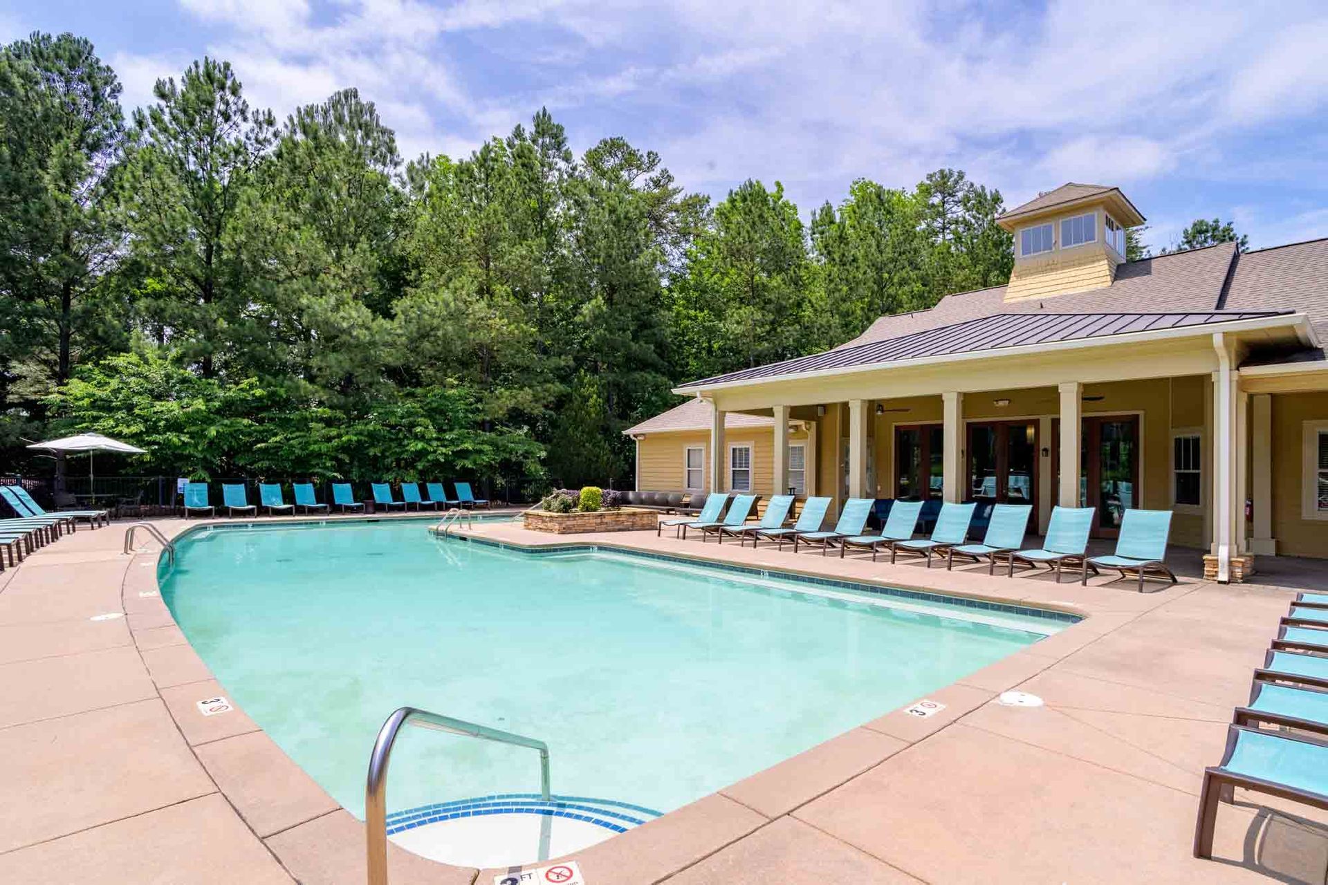 Swimming pool with blue lounge chairs, next to a building and trees on a sunny day.