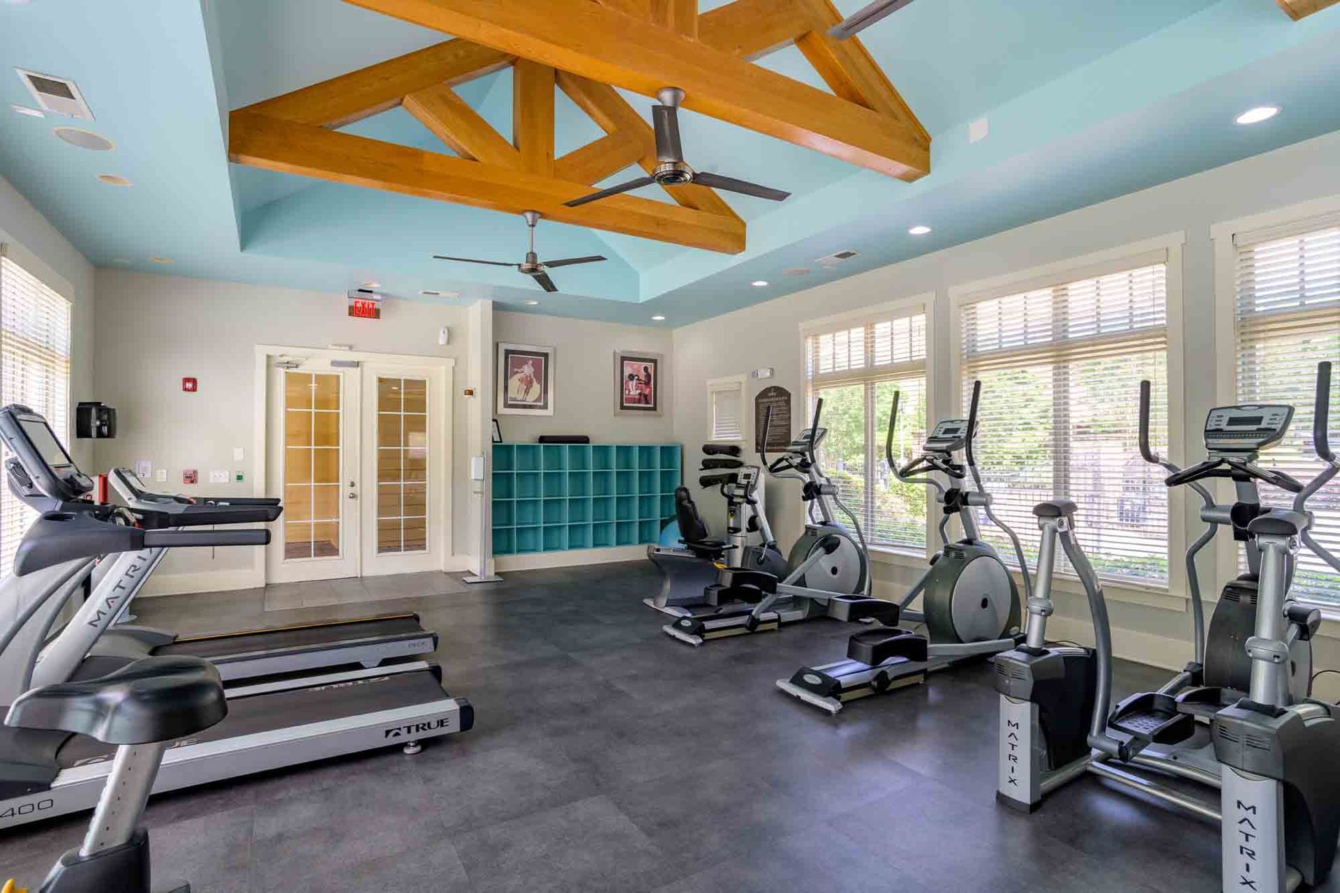 Gym with exercise equipment: treadmills, ellipticals, recumbent bikes. Blue ceiling with wooden beams.