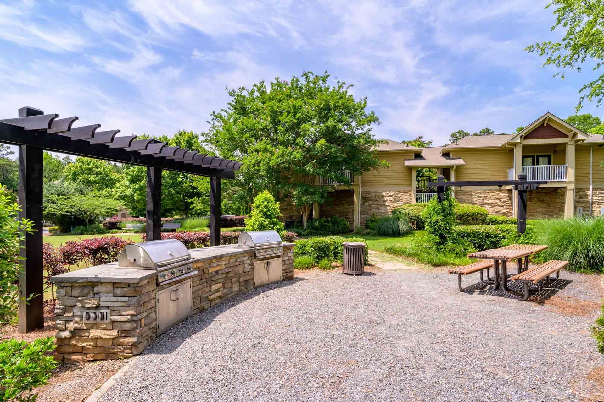 Outdoor grilling area with stone grill, pergola, picnic table, and apartment building in background.