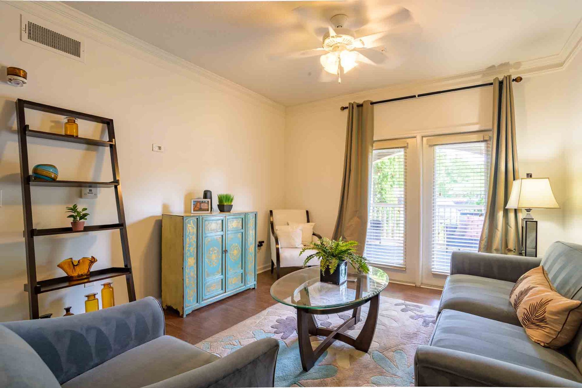 Living room: teal cabinet, gray sofa, glass table, wooden shelves, balcony access, tan curtains.