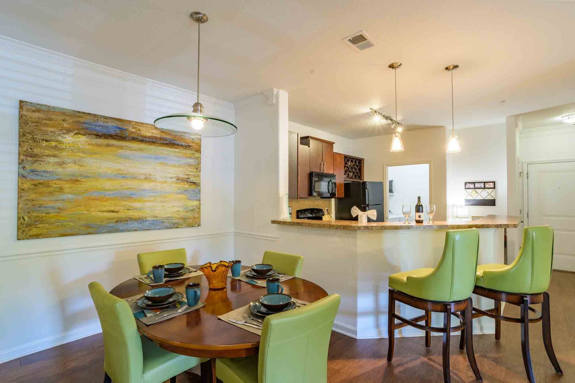 Dining room with round table, green chairs, and abstract art; kitchen with bar stools.